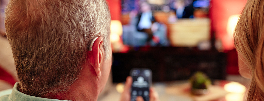 Image of male hearing aid wearer and female companion watching TV and using mobile phone to control volume